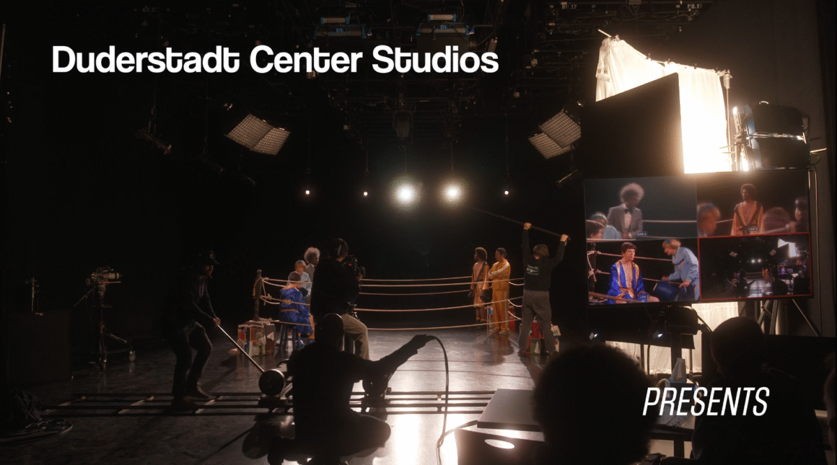 Film studio scene with crew members filming actors inside a boxing ring under stage lights. On-screen text reads “Duderstadt Center Studios” and “Presents.”