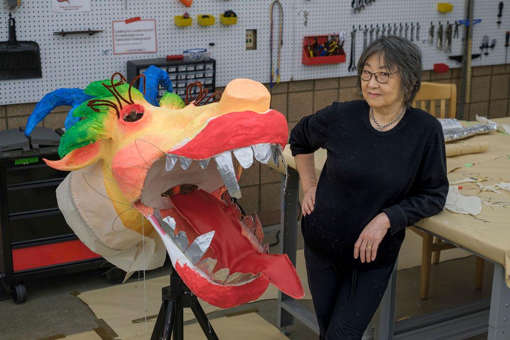 Artist Zhen Guo stands beside a colorful dragon head sculpture made from recycled materials in the Duderstadt Center Design Lab workshop.