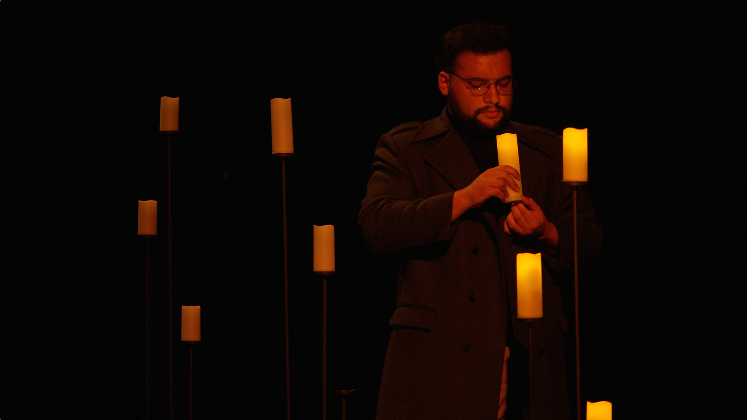 Person standing on a dark stage with candles around him.
