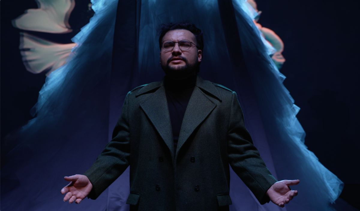 Man on stage with a mustache and military style jacket with a flowing skirt behind him. Both are in blue light.