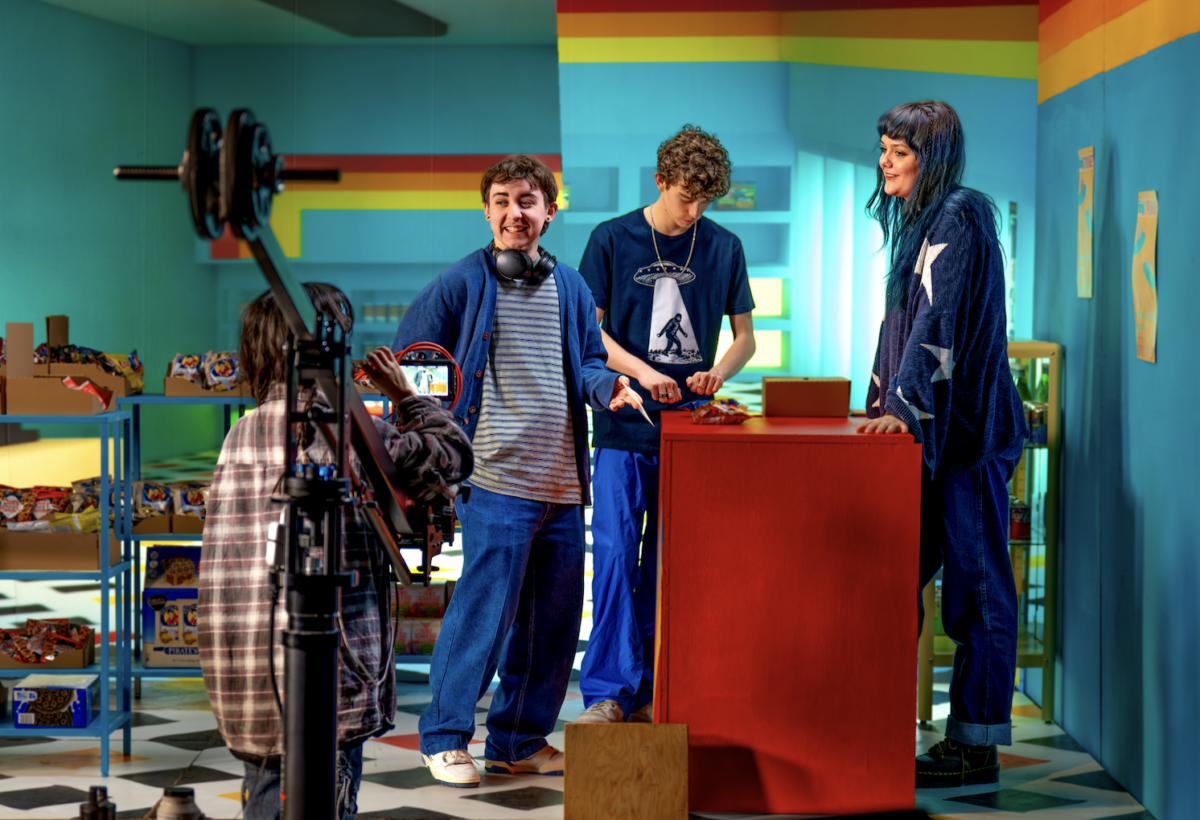 3 student actors on a blue set with a red table and a camera person filming them.